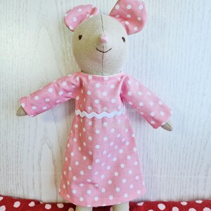 May include: A stuffed mouse toy with beige fabric skin, wearing a pink dress with white polka dots and a white rickrack trim at the waist. The mouse has large pink ears with white polka dots, embroidered dark eyes, and a stitched nose and mouth.