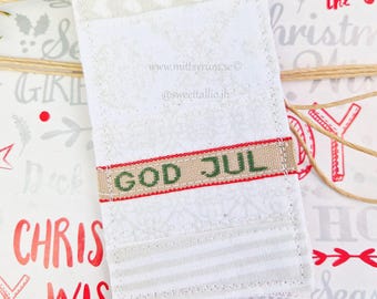 May include: A white fabric gift tag with a red and brown ribbon and the text "GOD JUL" in green letters. The tag is decorated with a white and gray pattern and has the website address "www.mittsyrum.se" and the social media handle "@sweettallio.jh" printed on it.
