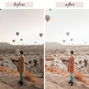 May include: A person in a brown coat stands on a rocky hilltop overlooking a valley with hot air balloons in the sky. The sky is a light blue with a few clouds.