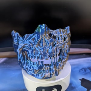 May include: A 3D printed half-face mask featuring a detailed skeletal jaw and nose structure. The mask has a metallic, iridescent finish that shifts between shades of blue, purple, and bronze, with visible horizontal layer lines from the printing process.