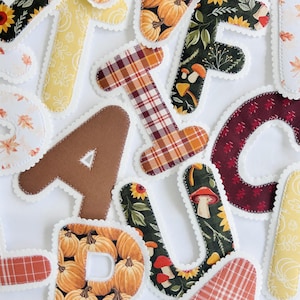 May include: A collection of fabric letters featuring various autumn-themed patterns including pumpkins, mushrooms, sunflowers, plaid, and solid brown colors. Each letter has a white scalloped edge finish, creating a decorative set of alphabet characters.