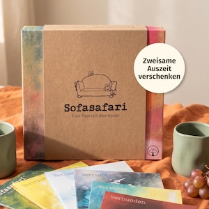 May include: A brown gift box with the text "Sofasafari" and "Euer Paarzeit Abenteuer" printed on the front. The box has a colourful design on the sides. A white circle with the text "Zweisame Auszeit verschenken" is on the right. Several colourful cards are in front.