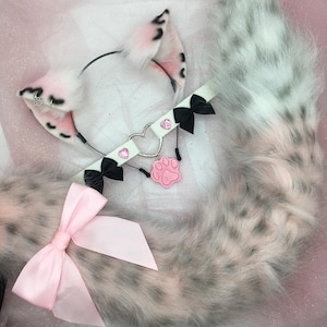 May include: A white and gray faux fur tail, headband with ears, and a choker set. The ears and tail have pink accents and black spots. The choker is white with black bows, a heart-shaped charm, and a pink paw print.