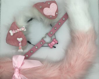 May include: A set of pink and white faux fur animal ears on a headband, a pink faux leather collar with rhinestones and a heart-shaped ring, and a long pink and white faux fur tail accented with a pink satin ribbon bow and bone-shaped charms.