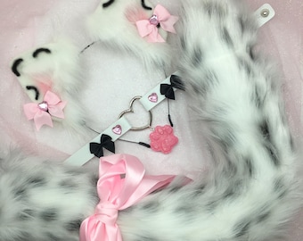 Puede incluir: Cola de piel sintética blanca con manchas negras, un par de orejas de gato de piel sintética blanca a juego con lazos rosas y un collar negro y blanco con un dije de corazón y un dije de pata rosa.