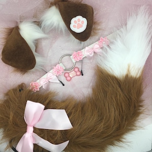 May include: A brown and white dog ear headband, tail, and collar set. The ears have a pink paw print and the collar has pink bows and flowers. The tail is brown and white, and the collar has a pink bone charm.