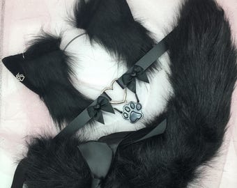 May include: Black faux fur cat ears headband with a black choker and a silver charm with a paw print. The choker has black satin ribbon bows.