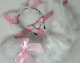 Pode incluir: Um conjunto com uma tiara de orelhas de gato em pelo sintético branco e rosa com corações prateados, uma gargantilha rosa com um pendente de coração e pata, e uma cauda longa de pelo sintético branco com um laço de cetim rosa claro.