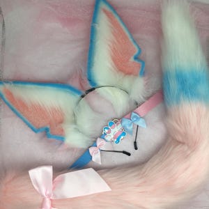 May include: A fluffy, pastel-colored cosplay set featuring a headband with ears, a tail, and a choker. The ears and tail have white, pink, and blue accents. The choker has a paw print charm and small bows. The tail is long and fluffy.