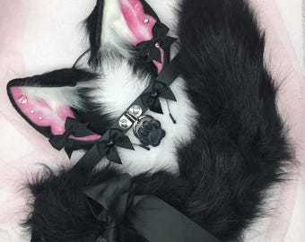 May include: Black and white faux fur cat ears with pink accents and a black choker with a paw charm. The ears have black bows and the choker has black bows and a silver chain.