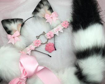 Op de afbeelding: Een wit-zwarte kattenstaart met een roze strik, een wit hoofdband met roze bloemen en een pootbedel, en een paar wit-zwarte kattenoren met roze strikken.