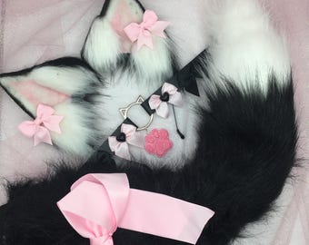 Puede incluir: Una cola esponjosa de color blanco y negro con un lazo rosa, un par de orejas de gato esponjosas de color blanco y rosa con lazos rosas y un dije de huella de pata blanco y negro.