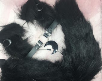 May include: A black faux fur tail, ears, and choker set. The choker has a silver ring and a black crescent moon charm. The word "OUJIA" is printed on the choker. The ears have silver hoops.