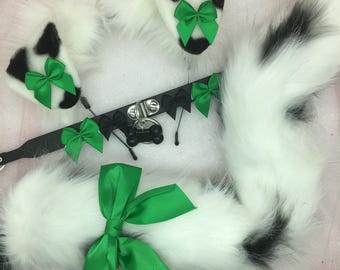 May include: A white and black faux fur tail with a black leather collar and green bows. The collar has a metal ring and a metal bone charm.
