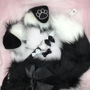 May include: A black and white faux fur tail with a white faux fur collar and a black heart-shaped charm with a bone pendant. The collar has a black bow and a white bow.