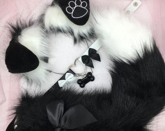 Puede incluir: Una cola de piel sintética negra y blanca con un collar de piel sintética blanca y un dije en forma de corazón negro con un dije de hueso. El collar tiene un lazo negro y un lazo blanco.