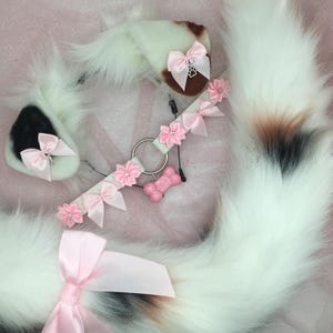 May include: A white and brown faux fur cat tail and ear set with pink bows and a matching collar. The collar features pink flower accents and a pink bone charm. The ears have black and brown accents.