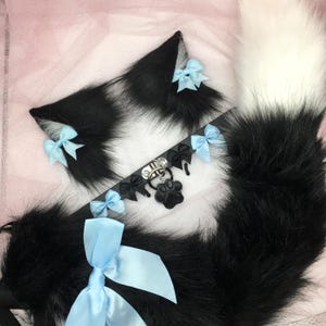 May include: Black and white faux fur cat ears with light blue bows, a black faux fur tail with a light blue bow, and a black choker with a paw charm and light blue bows.