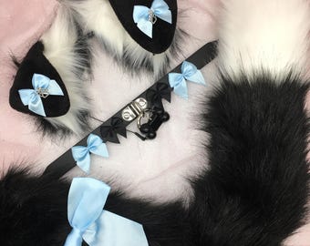May include: A black and white faux fur tail, ears, and collar set. The ears and collar feature light blue bows and black accents. The collar includes a bone-shaped charm. The tail is long and fluffy.