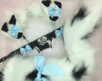 May include: A white and black faux fur cosplay set featuring a tail, ears, and a collar. The ears and collar have light blue bows. The collar includes a bone-shaped tag. The tail has black spots.