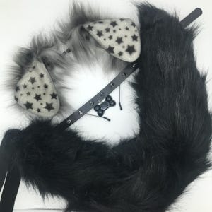 May include: A set of faux fur animal ears with a cream-colored base and black star patterns, featuring long grey fur trim. A black faux leather collar with silver-toned metal studs and a black bone-shaped charm, alongside a long black faux fur tail.