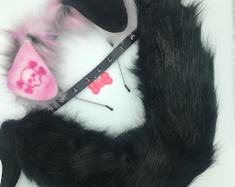 May include: A set of faux fur animal ears on a headband, a black studded collar with a pink bone-shaped charm, and a long black faux fur tail. The ears feature a pink and white color scheme with a prominent pink skull and crossbones graphic.