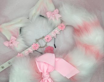Puede incluir: Una cola de gato de piel sintética rosa y blanca con un lazo de satén rosa, una diadema de piel sintética blanca con orejas de gato rosas y un lazo de satén rosa, y un collar de cuero blanco con abalorios de flores rosas.