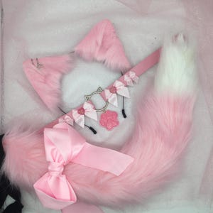 May include: A pink faux fur cat ear headband, collar, and tail set. The ears have silver hoops and the collar features a cat-ear charm, pink bows, and a paw print pendant. The tail is pink with a white tip and a large pink bow.