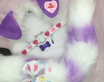 May include: A white and purple faux fur tail with a purple bow and a white collar with pink hearts and the text "Be Mine".