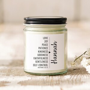 May include: A white candle jar with a black lid and a white label. The label has the text "LOVE, JOY, PEACE, PATIENCE, KINDNESS, GOODNESS, FAITHFULNESS, GENTLENESS, SELF-CONTROL, GALATIANS 5:22-23" and the name "Hannah" written vertically.