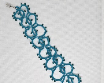 May include: A teal tatting lace bracelet featuring a series of interconnected circular and petal-shaped motifs. The lace is embellished with small, matching teal beads along the edges and is finished with silver-toned metal end caps and a clasp.