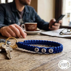 May include: A blue braided keychain wrist strap with the letters "PSG" in white beads. The keychain is attached to a gold-colored ring and a set of keys. The background includes a coffee cup and a person using a phone.