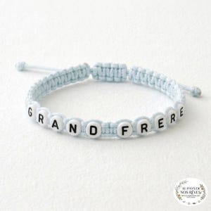 Puede incluir: Una pulsera de macramé azul claro con cuentas blancas con letras que deletrean "GRAND FRERE". La pulsera es ajustable y tiene un cierre de nudo corredizo. Es un accesorio personalizado.