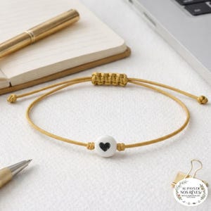 Può includere: Braccialetto regolabile giallo senape con una perla bianca e un cuore nero. Il braccialetto è realizzato con un cordoncino intrecciato. È esposto su una superficie bianca con una penna, un taccuino e un laptop sullo sfondo.