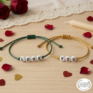 May include: Two friendship bracelets, one green and one gold, each with white letter beads spelling "LOVE" and "YOU". The bracelets are on a wooden surface with rose petals and a candle.