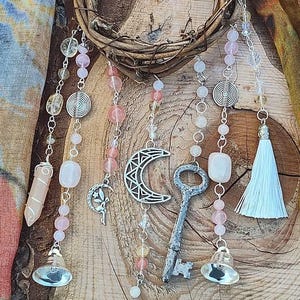 May include: A wooden wreath with pink and clear crystal beads, a silver key, a crescent moon charm, and a white tassel hanging from it.
