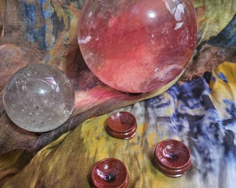 May include: Two crystal spheres, one large and reddish-pink, the other smaller and clear, rest on a colorful fabric. Three small, round, wooden stands are also present. The spheres appear to be made of quartz.