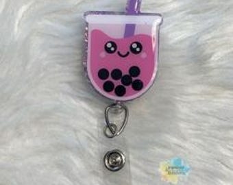 May include: A pink and purple retractable badge reel in the shape of a boba tea cup. The cup has a smiling face, black boba pearls, and a purple straw. The reel has a silver clip and a clear plastic strap.