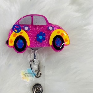 May include: A pink glittery car-shaped badge reel with yellow and blue flower accents and a peace sign. The badge reel has a clear plastic holder and a retractable cord. The text "Impulsive Kreations" is visible on the badge reel.