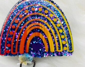 May include: A badge reel with a rainbow design. The rainbow features layers of orange, yellow, and blue glitter. The badge reel has a retractable cord and a metal clip. The text "Impulsive Creations" is visible on the badge reel.