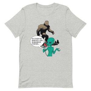 May include: A grey t-shirt with a cartoon illustration of a green alien on the phone with a brown Bigfoot. The alien is saying "So I know you guys will like never believe this but..."