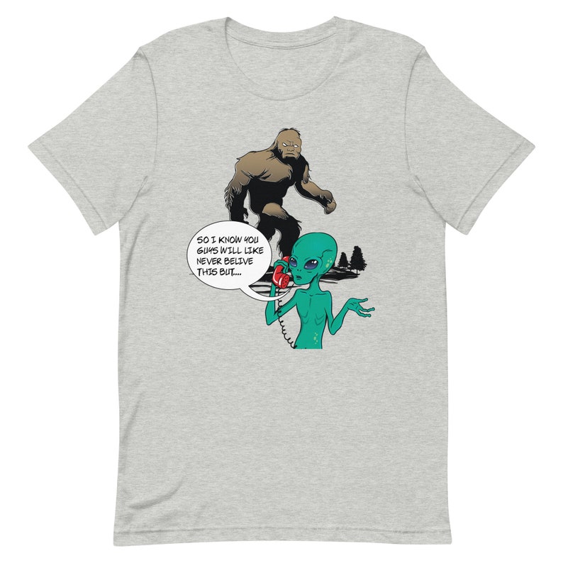 May include: A grey t-shirt with a cartoon illustration of a green alien on the phone with a brown Bigfoot. The alien is saying "So I know you guys will like never believe this but..."