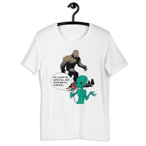 May include: A white t-shirt with a green alien on the phone with a brown Bigfoot. The alien is saying "So I know you guys will like never believe this but..."