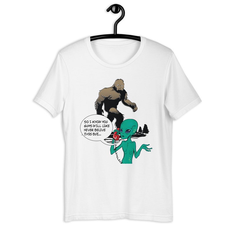 May include: A white t-shirt with a green alien on the phone with a brown Bigfoot. The alien is saying "So I know you guys will like never believe this but..."