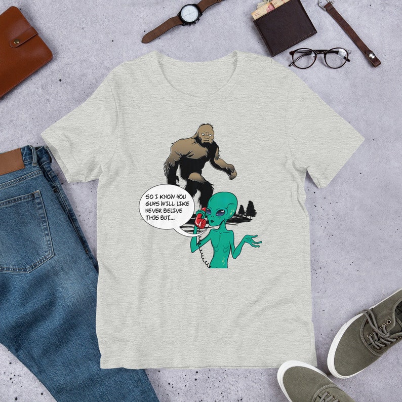 May include: A gray t-shirt with a cartoon illustration of a Bigfoot talking on the phone to an alien. The text on the shirt reads "So I know you guys will like never believe this but..."
