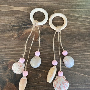 May include: Two wooden rings with twine hanging from them. Each ring has three strands of twine with a pink bead, a seashell, and another pink bead. The seashells are different shapes and sizes.