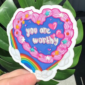 Pode incluir: Um adesivo em forma de cora&ccedil;&atilde;o com um arco-&iacute;ris e nuvens. O adesivo tem cobertura rosa com granulado branco e estrelas. O texto no adesivo diz "You are worthy".