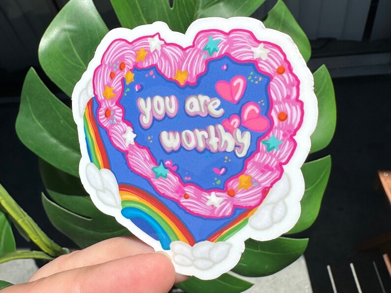 Pode incluir: Um adesivo em forma de cora&ccedil;&atilde;o com um arco-&iacute;ris e nuvens. O adesivo tem cobertura rosa com granulado branco e estrelas. O texto no adesivo diz "You are worthy".