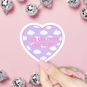 May include: A heart-shaped sticker with a purple background and white clouds. The sticker says "i am the main character" in pink letters.