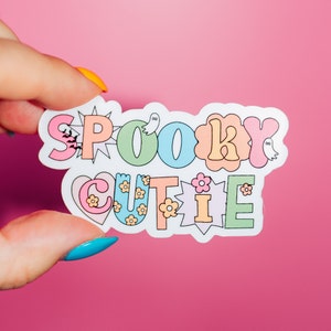 May include: A colourful sticker with the words "SPOOKY CUTIE" in a retro font. The sticker features bats, ghosts, and flowers. The background is a vibrant pink.